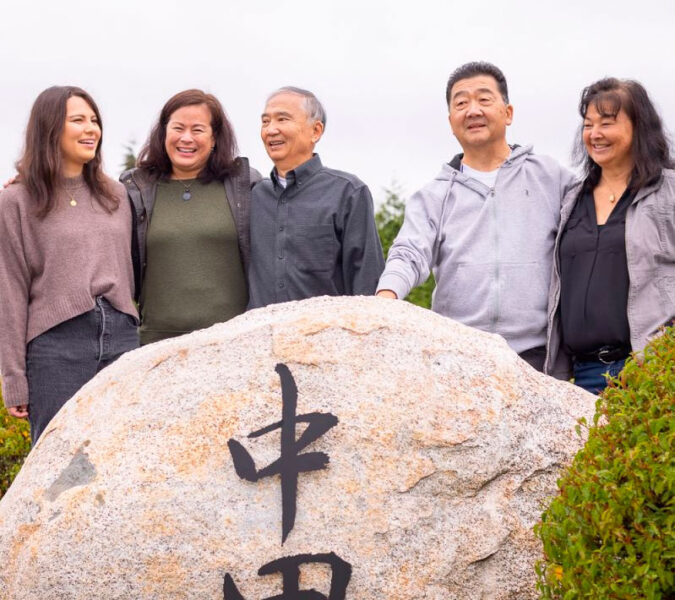 Five adults smiling and posing behind a large rock with Chinese characters outdoors.