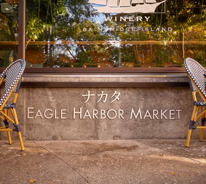Storefront signage reading 'Eagle Harbor Market' with Japanese characters, framed by outdoor seating on a sidewalk in front of a winery window reflection.
