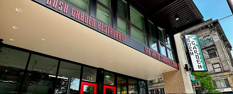 Front view of Bush Garden Restaurant with red doors and large glass storefronts on a sunny street corner.