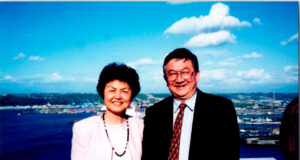 Smiling older couple posing on a ship's railing with a harbor and blue sky in the background.