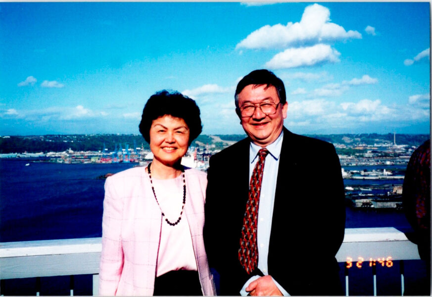 Smiling older couple posing on a ship's railing with a harbor and blue sky in the background.