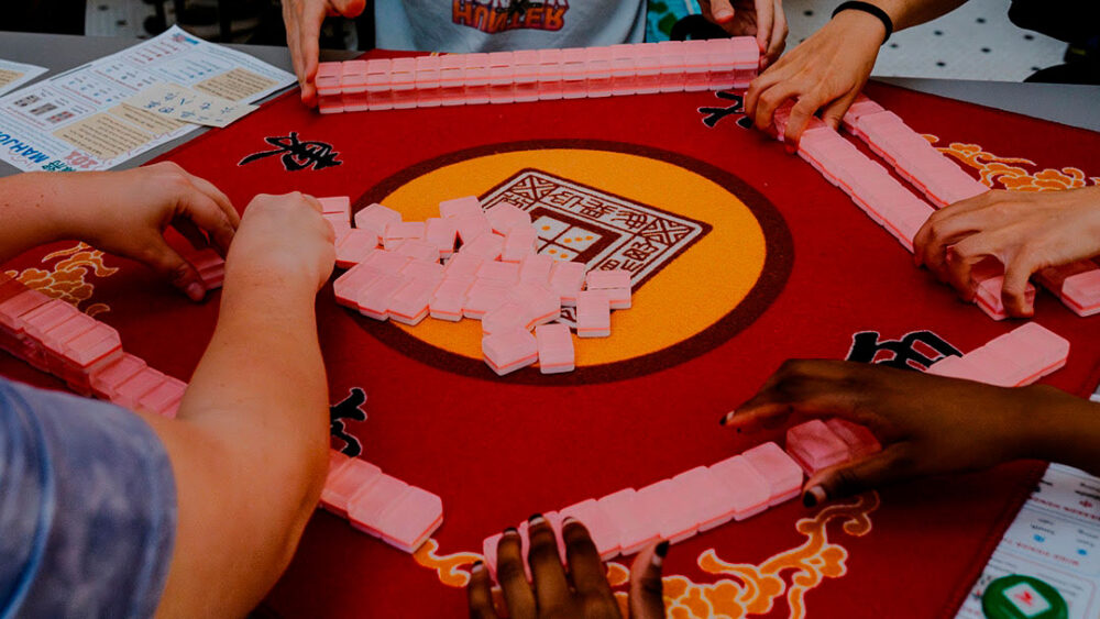 People playing a pink-tiled Mahjong game around a red circular table, hands actively arranging tiles.