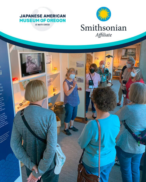 Group of visitors listening to a guide in a museum exhibit, all wearing masks, with display cases and artifacts around them.