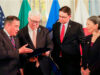 Four adults in suits review a folder or binder during a formal ceremony, with national flags in the background.