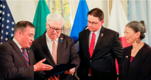 Four adults in suits review a folder or binder during a formal ceremony, with national flags in the background.