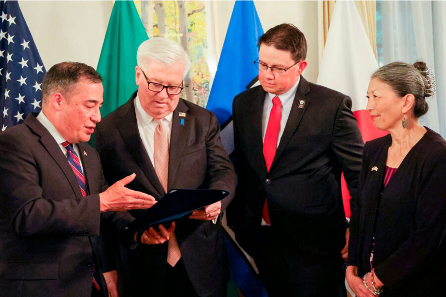 Four adults in suits review a folder or binder during a formal ceremony, with national flags in the background.