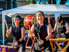 Three taiko drummers in black outfits perform under a white canopy, with a large drum in the foreground and wooden stands nearby.