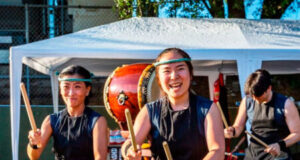 Three taiko drummers in black outfits perform under a white canopy, with a large drum in the foreground and wooden stands nearby.