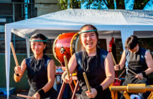 Three taiko drummers in black outfits perform under a white canopy, with a large drum in the foreground and wooden stands nearby.