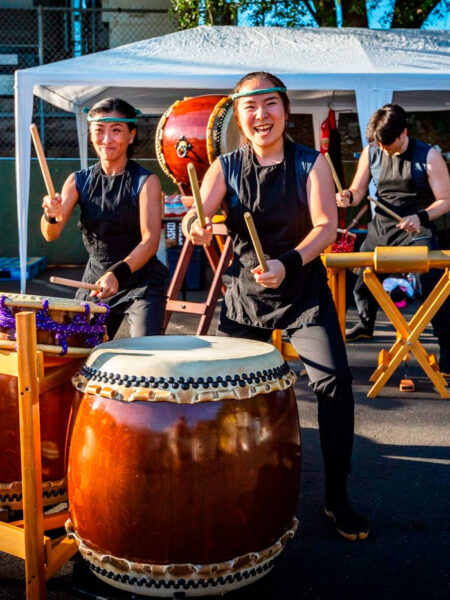 Three taiko drummers in black outfits perform under a white canopy, with a large drum in the foreground and wooden stands nearby.