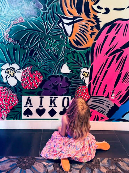 Girl in a floral dress sits on a dark tile floor in front of a bright mural with a tiger, tropical plants, strawberries, and the word AIKO on a sign.