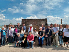 Group of about 20 people posing in front of a stone monument outdoors on a sunny day
