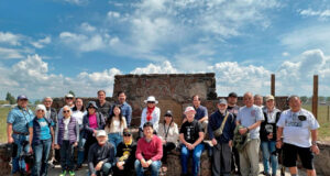 Group of about 20 people posing in front of a stone monument outdoors on a sunny day