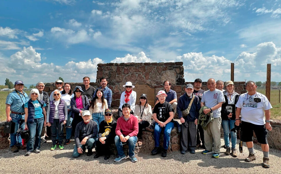 Group of about 20 people posing in front of a stone monument outdoors on a sunny day