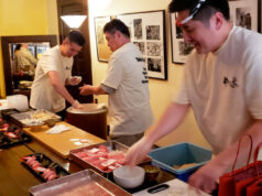Three chefs in white shirts prepare sushi at a busy counter with trays of fish and bowls nearby.