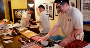 Three chefs in white shirts prepare sushi at a busy counter with trays of fish and bowls nearby.