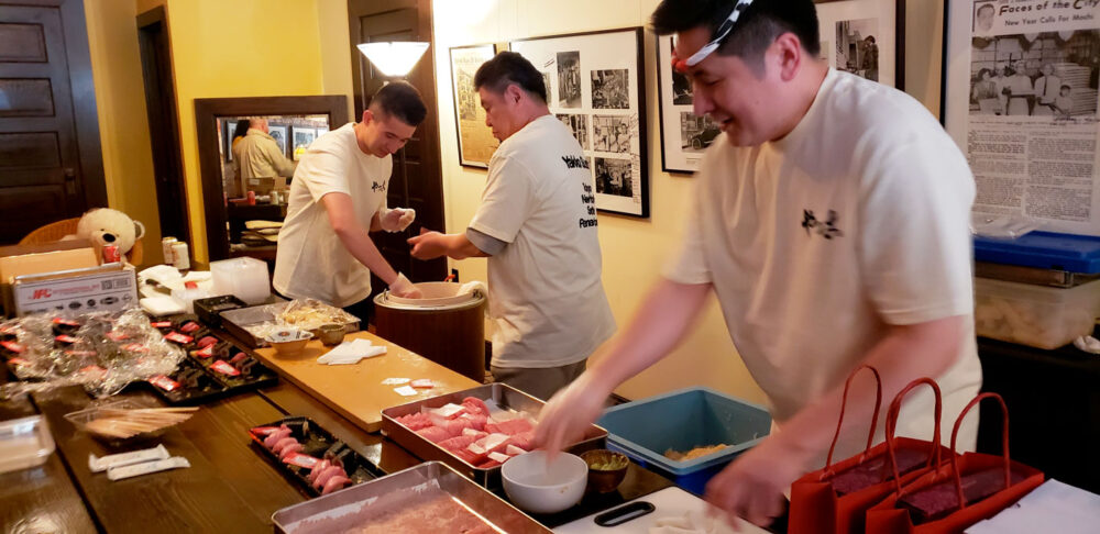 Panama-3-chefs---20260507_180720 | Seattle’s Japanese Community News – North American Post Three chefs in white shirts prepare sushi at a busy counter with trays of fish and bowls nearby.