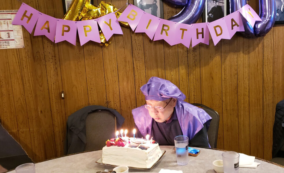 Person in a purple hat leaning toward a frosted birthday cake with lit candles at a table, pink'HAPPY BIRTHDAY' banner overhead.