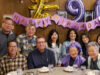 Happy 90th Birthday, Tomio Group of friends and family posing at a birthday party around a table with a 'Happy Birthday' banner and purple balloons in the background.