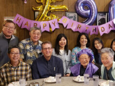 Group of friends and family posing at a birthday party around a table with a 'Happy Birthday' banner and purple balloons in the background.