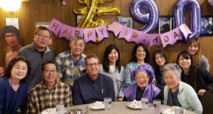 Group of friends and family posing at a birthday party around a table with a 'Happy Birthday' banner and purple balloons in the background.