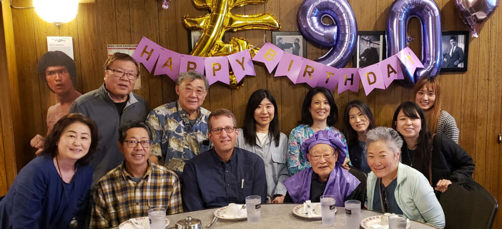 Group of friends and family posing at a birthday party around a table with a'Happy Birthday' banner and purple balloons in the background.