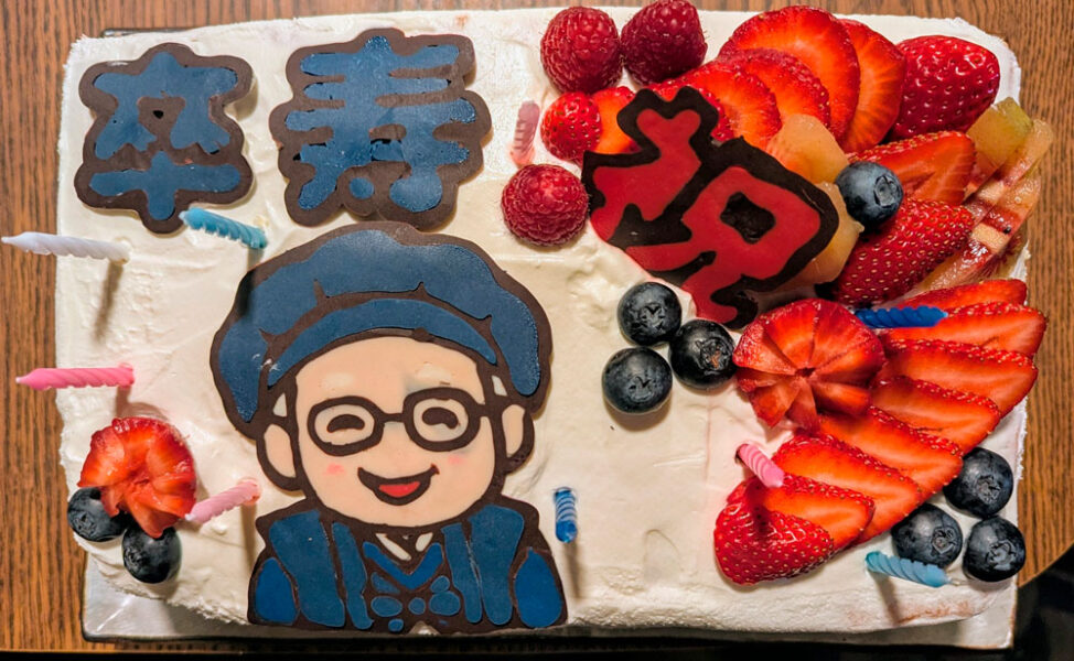 White frosted birthday cake with a cartoon person in a blue hat and glasses, surrounded by strawberries, blueberries, raspberries, and candles, plus blue and red Chinese characters on top.