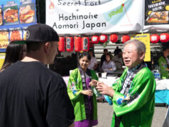 Festival scene with people in green traditional jackets at a Japanese food stall decorated with red lanterns and banners advertising Hachinohe Aomori Japan on a sunny day (informative)