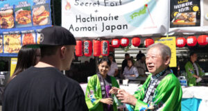 Festival scene with people in green traditional jackets at a Japanese food stall decorated with red lanterns and banners advertising Hachinohe Aomori Japan on a sunny day (informative)
