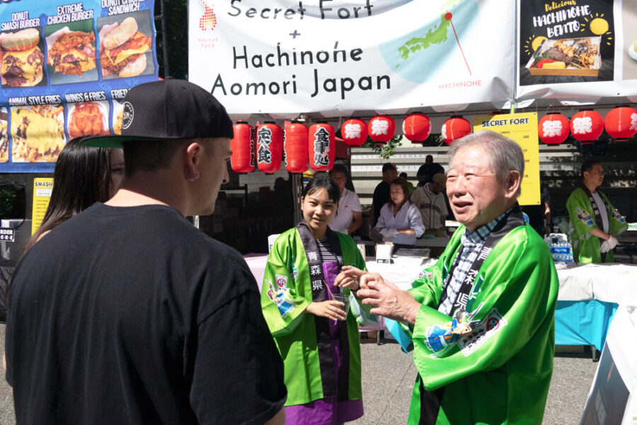 _-main | Seattle’s Japanese Community News – North American Post Festival scene with people in green traditional jackets at a Japanese food stall decorated with red lanterns and banners advertising Hachinohe Aomori Japan on a sunny day (informative)