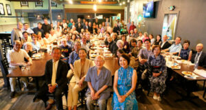 Large group photo of diverse people seated and standing around long restaurant tables for a group portrait.