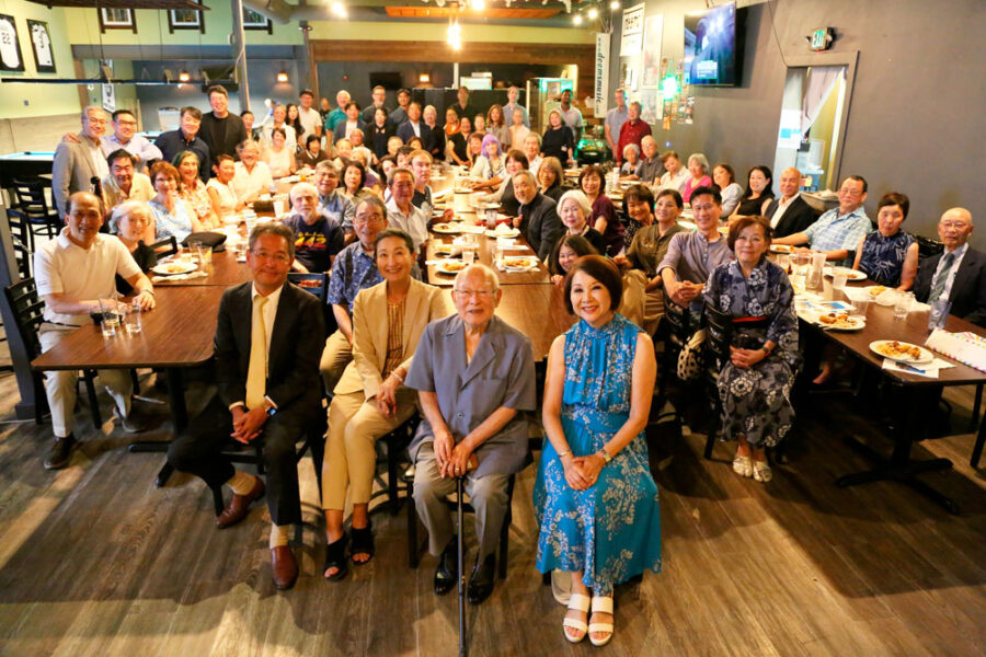 Large group photo of diverse people seated and standing around long restaurant tables for a group portrait.