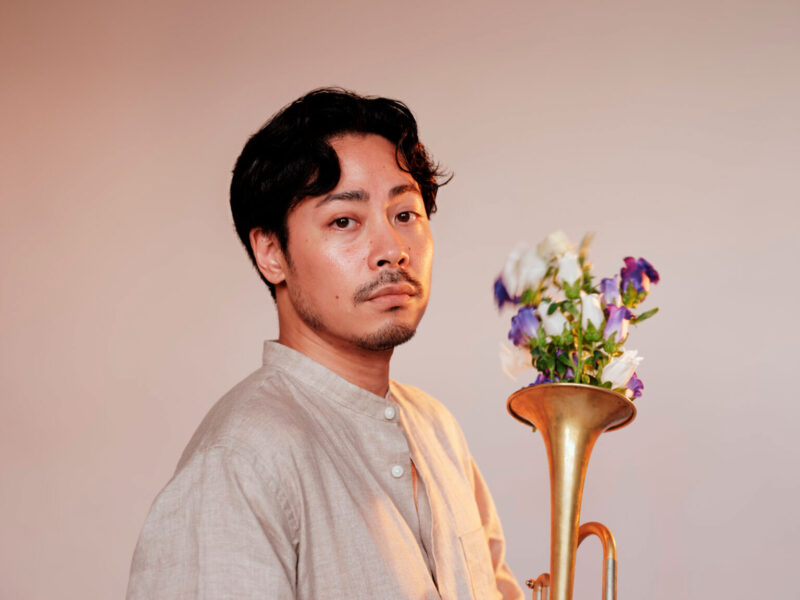 Portrait of a man in a light linen shirt beside a brass instrument with a bouquet of purple and white flowers in its bell]","A man looks at the camera next to a brass instrument filled with purple and white flowers]","Gentleman posing with a brass horn and a flower arrangement in the bell, neutral background]","Informational: man posing with brass instrument and floral bouquet for a musical-themed portrait]","Close-up portrait of a man with a brass instrument and a flower bouquet inside the bell]