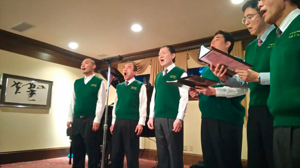 Group of men in green vests sing on a stage, holding music folders and a microphone in front of them