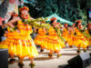 Event Calendar From August 28th Print Isuue Group of women in bright yellow dresses dancing with leafy garlands at an outdoor festival stage reading scene clearly as a hula performance.