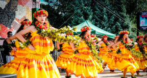 Group of women in bright yellow dresses dancing with leafy garlands at an outdoor festival stage reading scene clearly as a hula performance.