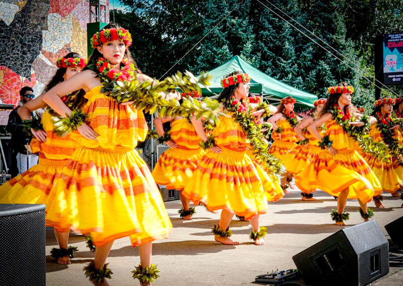 Group of women in bright yellow dresses dancing with leafy garlands at an outdoor festival stage reading scene clearly as a hula performance.
