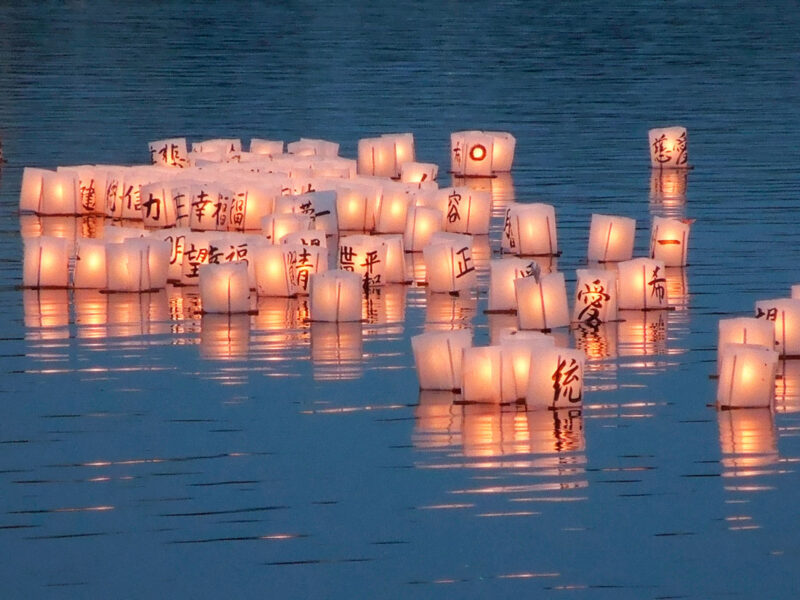 Glowing paper lanterns with Asian writing drift on a calm blue lake at night.