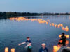 People wading in a lake at dusk, releasing glowing paper lanterns along the water, with a wooden dock in the foreground.