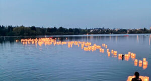 People wading in a lake at dusk, releasing glowing paper lanterns along the water, with a wooden dock in the foreground.