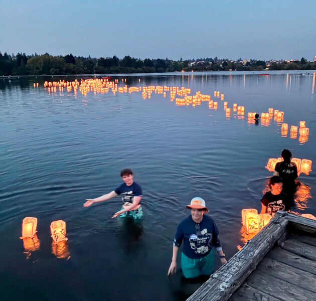People wading in a lake at dusk, releasing glowing paper lanterns along the water, with a wooden dock in the foreground.