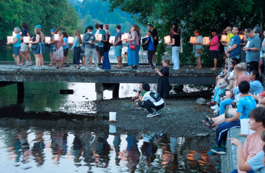 Crowd of people on a wooden bridge holding illuminated lanterns over a river at dusk, with photographers crouching below.
