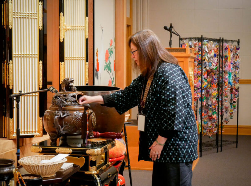 _-Interfaith-Karyn---20260802_BetsuinMemorialSvc_27 | Seattle’s Japanese Community News – North American Post Woman with a lanyard stands beside a large ornate bronze ceremonial basin on a decorative stand at a cultural display.