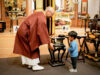 Buddhist monk in maroon robes guiding a young boy as they place incense in a ceremonial burner in a temple altar room.