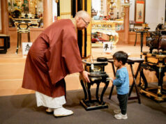 Buddhist monk in maroon robes guiding a young boy as they place incense in a ceremonial burner in a temple altar room.