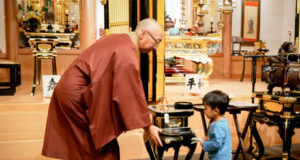 Buddhist monk in maroon robes guiding a young boy as they place incense in a ceremonial burner in a temple altar room.