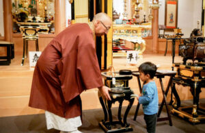 Hiroshima/Nagasaki Atomic Bomb Victims’ Interfaith Memorial Service Buddhist monk in maroon robes guiding a young boy as they place incense in a ceremonial burner in a temple altar room.