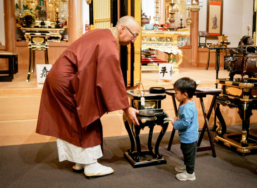 _-Interfaith-boy---20260802_BetsuinMemorialSvc_42 | Seattle’s Japanese Community News – North American Post Buddhist monk in maroon robes guiding a young boy as they place incense in a ceremonial burner in a temple altar room.