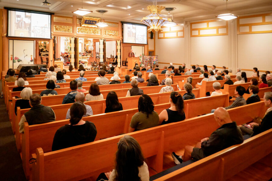 _-Interfaith-congregation---20260802_BetsuinMemorialSvc_32 | Seattle’s Japanese Community News – North American Post Audience seated in wooden pews facing a stage with a speaker at a podium and ornate altar, flanked by two projection screens showing images.