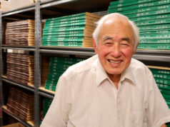 Smiling elderly man in a white shirt stands in a storage room with shelves of green and brown boxes.